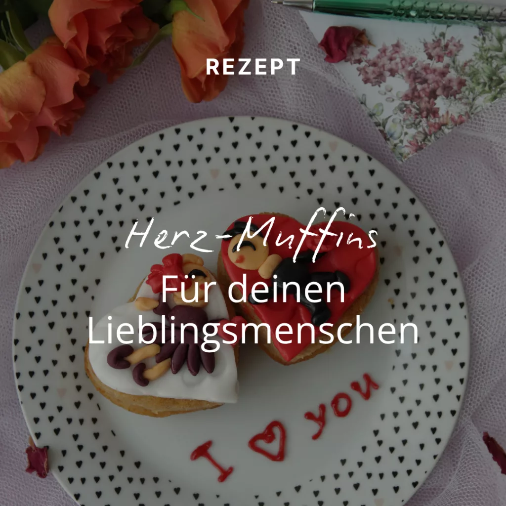 Ein Teller mit zwei verzierten herzförmigen Muffins, Blumen und einer Karte mit der Aufschrift „Ich liebe dich“. Der deutsche Text lautet: „Herz-Muffins für deinen Lieblingsmenschen“.