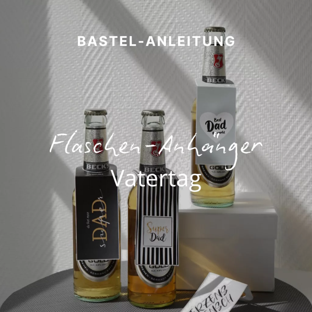 Auf einem Tisch stehen drei Bierflaschen mit Anhängern zum Vatertag. Auf den Anhängern steht „Der beste Papa aller Zeiten“, „Super-Papa“ und „Papa“. Das Sonnenlicht wirft Schatten an die Wand dahinter. Daneben steht eine kleine Schachtel mit einer Karte.