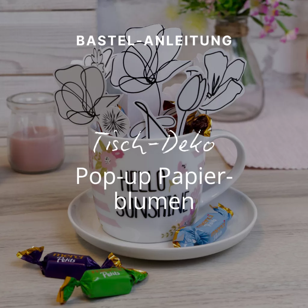 Eine Tasse mit ausgeschnittenen Papierblumen, die auf einer Untertasse mit Bonbons auf einem Holztisch steht. Über dem Bild ist ein deutscher Text eingeblendet, der eine Bastelanleitung für Pop-up-Papierblumen enthält.