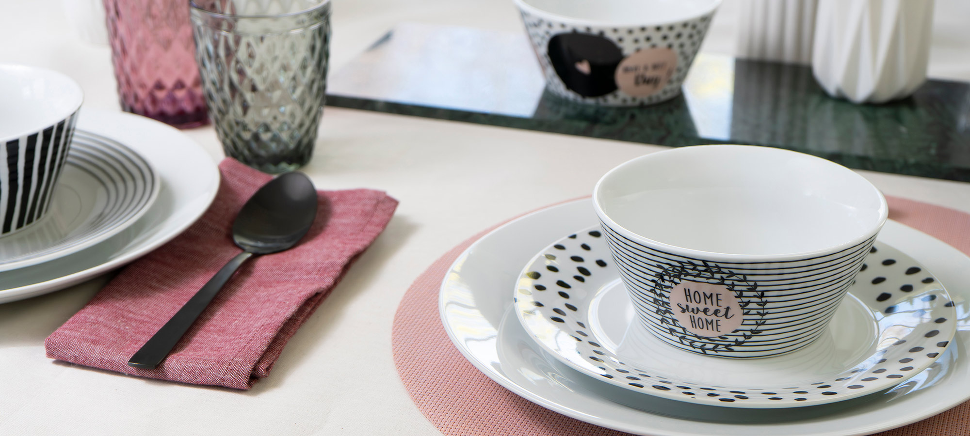 Ein gedeckter Tisch mit gemusterten Schalen, Tellern und Gläsern. Eine Schale mit der Aufschrift "Home sweet home" steht auf einem Teller mit einem rosa Tischset. Ein schwarzer Löffel ruht auf einer rosa Serviette. Im Hintergrund stehen dekorative Vasen und weiteres Geschirr.