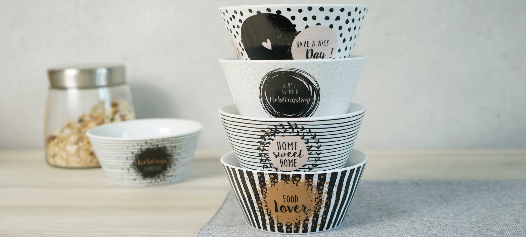 Vier dekorative Schalen mit verschiedenen schwarzen und weißen Mustern und positiven Botschaften sind auf einem Tisch gestapelt; im Hintergrund befinden sich eine Schale und ein mit Müsli gefülltes Glasgefäß.