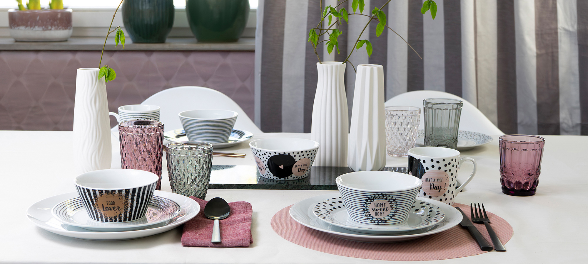 Ein Esstisch mit gemusterten Tellern, Schüsseln, Tassen und Gläsern in Weiß-, Rosa- und Grautönen. Weiße Vasen mit grünen Zweigen schmücken die Mitte. Utensilien und Servietten sind ordentlich angeordnet.