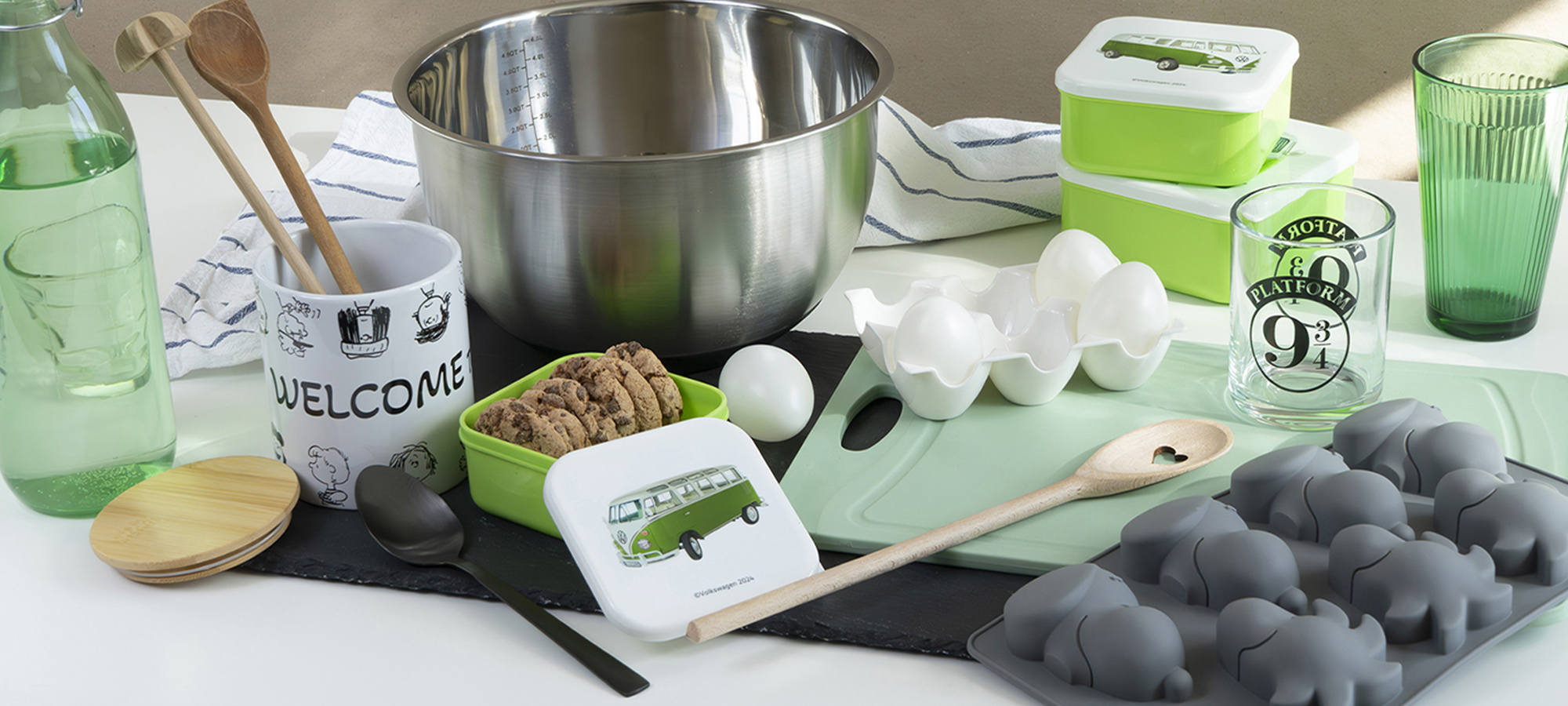 Auf einer Küchentheke stehen Backutensilien: eine Metallschüssel, Holzlöffel, Glaswaren, eine Wasserflasche, Becher, grüne und weiße Vorratsbehälter, Eier, ein Schneidebrett, Kekse in einer Dose und eine Eisschale in Elefantenform.