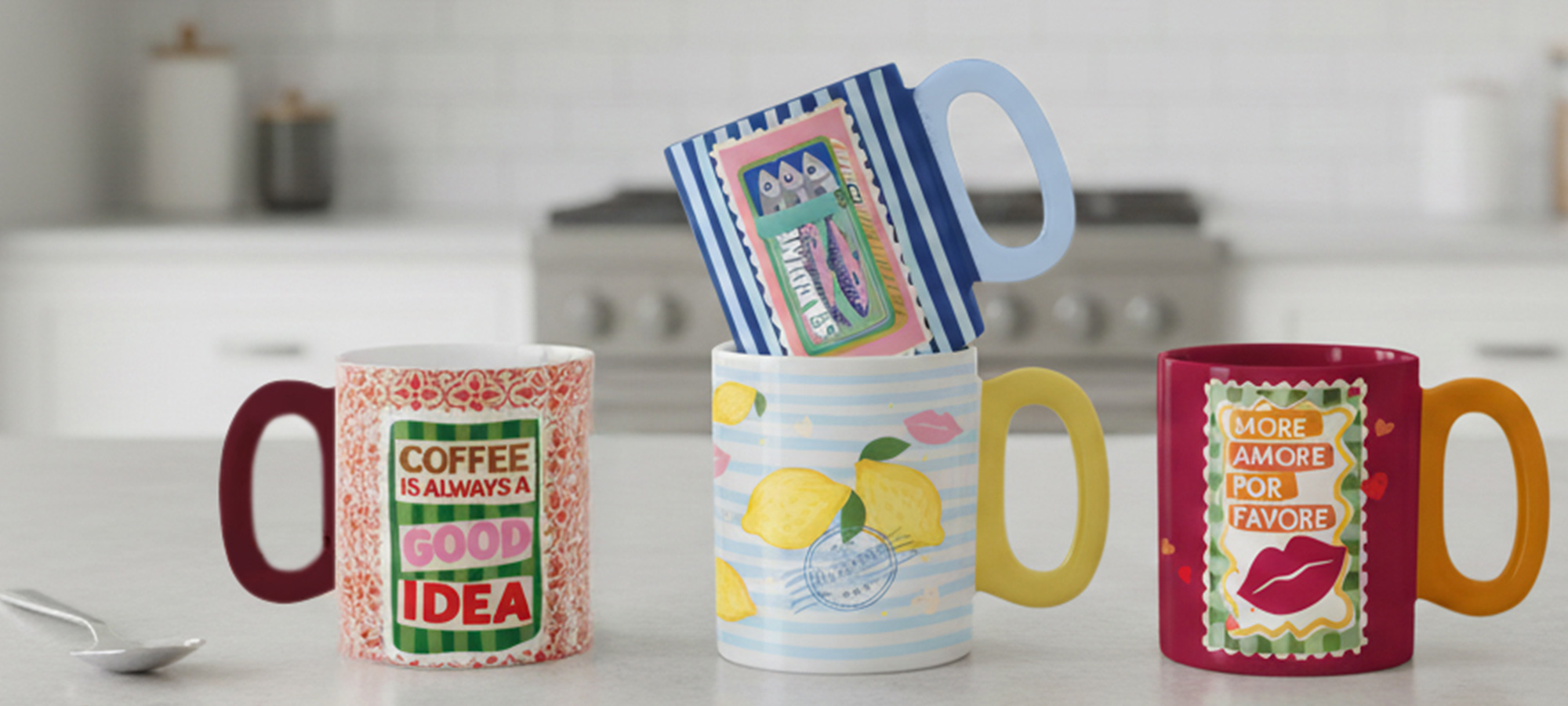 Drei bunte Tassen mit unterschiedlichen Mustern und Slogans stehen auf einem Küchentisch; eine Tasse wird waagerecht auf zwei aufrecht stehenden Tassen balanciert, und ein Löffel liegt daneben.