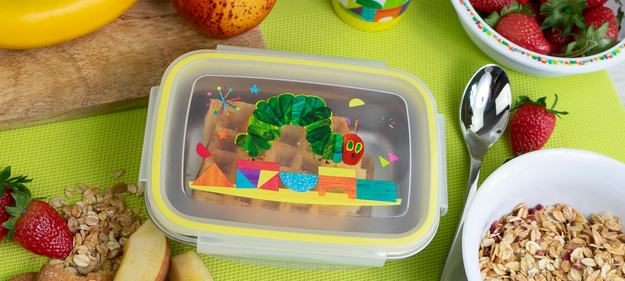 Auf einer grünen Matte steht ein Plastikbehälter mit dem Motiv der Raupe Nimmersatt, umgeben von Apfelscheiben, Erdbeeren, einer Schüssel mit Müsli, einem Löffel und Obst.