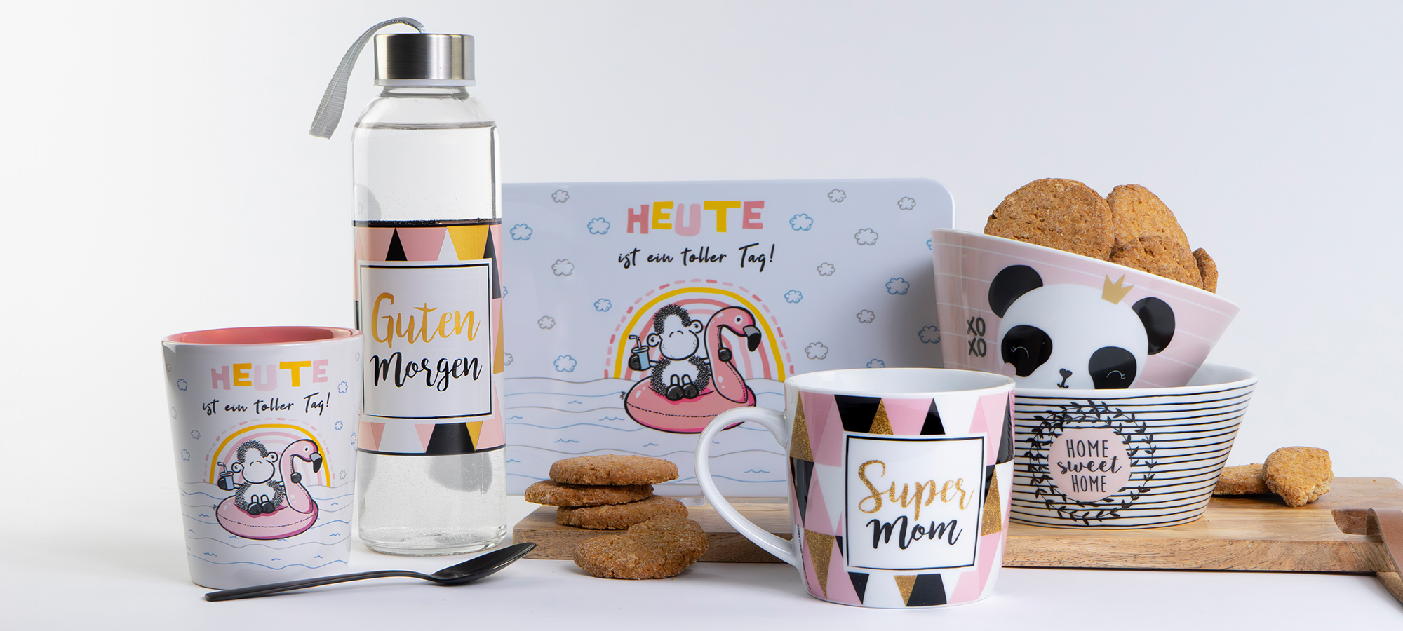 Eine Reihe von Tassen, eine Glasflasche, eine Schale mit Keksen, ein Tablett und eine Tasse, alle mit verspielten Designs und deutschen Sprüchen, angeordnet auf einem Holzbrett mit Keksen, vor einem weißen Hintergrund.