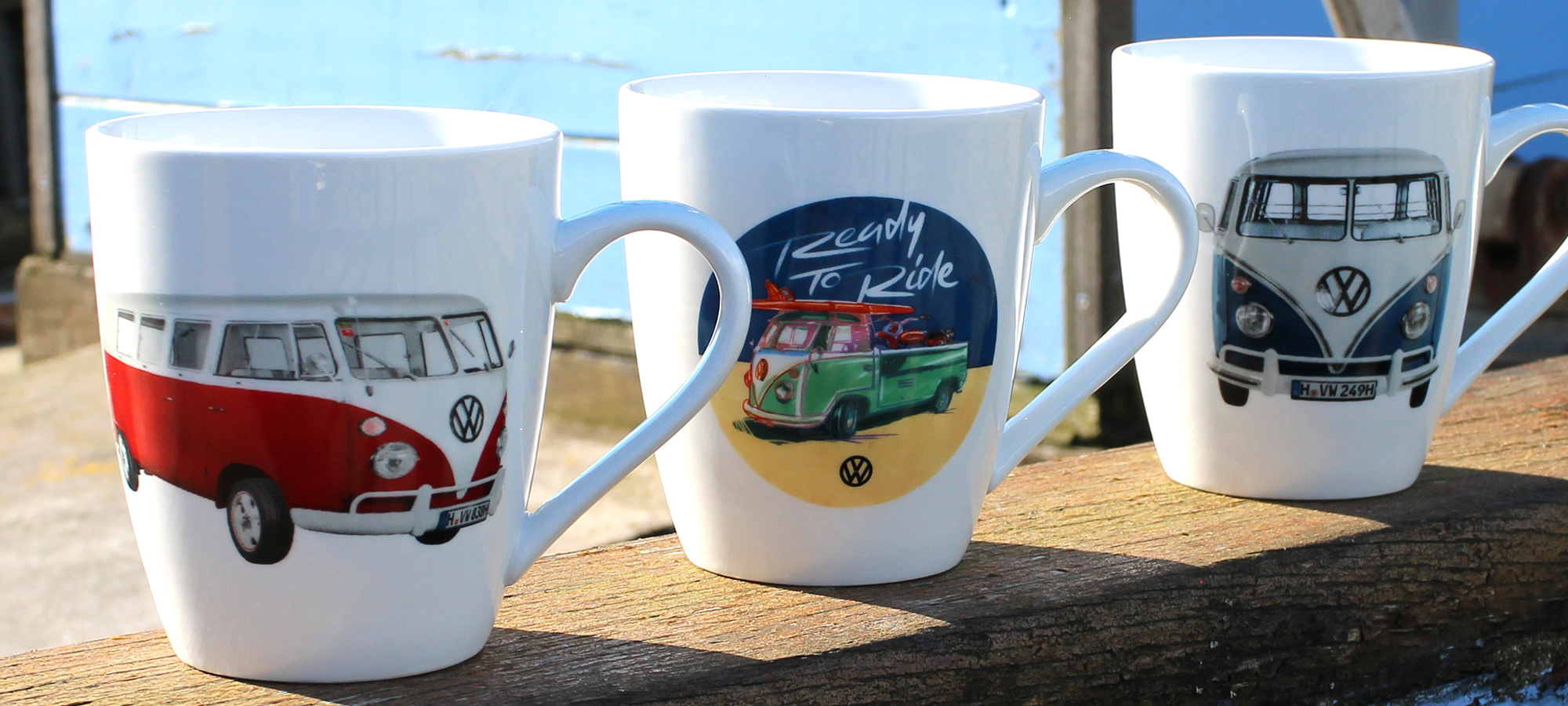 Drei weiße Tassen mit bunten Volkswagen-Wohnmobilmotiven und -Logos stehen auf einer sonnenbeschienenen Holzfläche mit einem verschwommenen blauen Hintergrund.