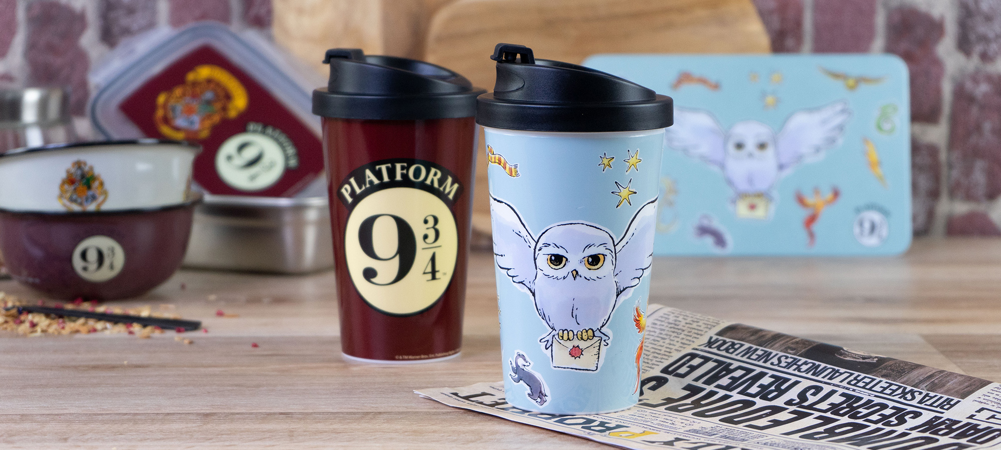Zwei Reisebecher auf einem Holztisch: ein roter mit einem Bahnsteig 9 3/4-Logo und ein hellblauer mit einer Cartoon-Eule und Hogwarts-Themen. Im Hintergrund sind Küchenutensilien und Dekoration zum Thema Harry Potter zu sehen.