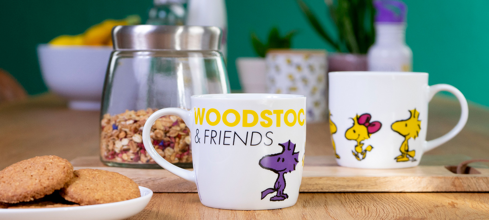 Ein weißer Becher mit Woodstock & Friends und einem Cartoon-Vogel steht auf einem Holztisch neben Keksen, Müsli im Glas und einem weiteren Becher mit gelben Vögeln vor einem verschwommenen grünen Hintergrund.