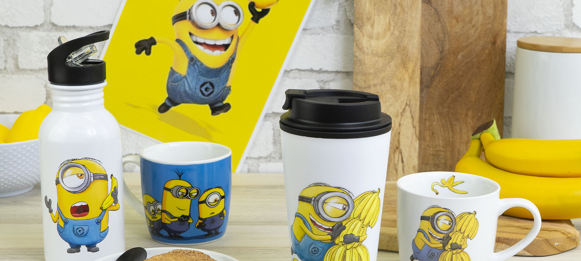 Eine Sammlung von Tassen, eine Wasserflasche und ein Reisebecher mit den Minions aus Despicable Me, die auf einem Tisch mit Bananen, Keksen und einem Minions-Poster im Hintergrund stehen.