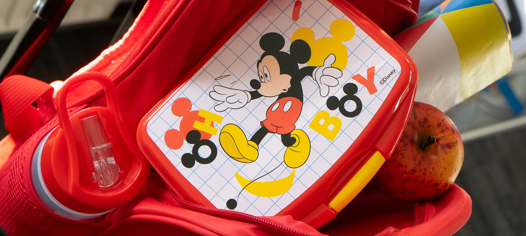 Eine rote Brotdose mit einer Zeichentrickfigur von Mickey Mouse, dem Wort BOY und bunten Formen befindet sich in einem roten Rucksack, in dem eine Wasserflasche und ein Apfel stehen.