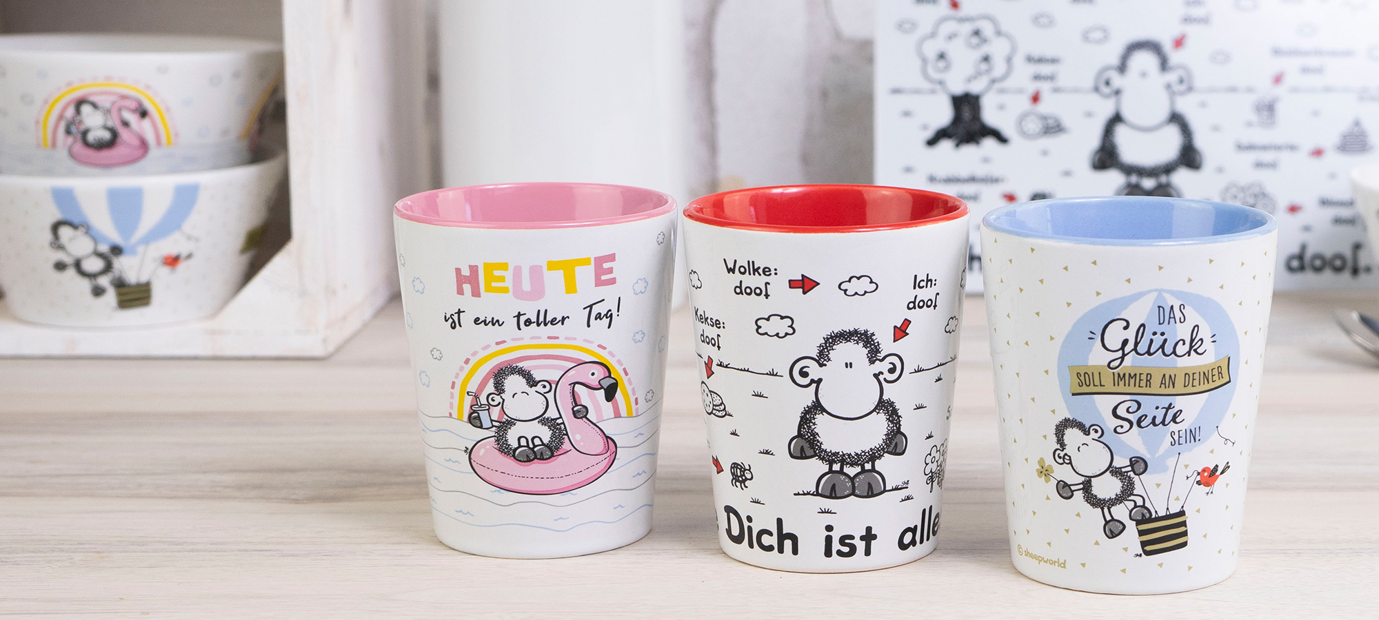 Drei illustrierte Tassen mit Schafsmotiven sind auf einem Tisch aufgereiht. Jede Tasse hat bunte Ränder und verschiedene deutsche Sprüche. Im Hintergrund befinden sich zwei Schalen und ein dekoratives Brett mit ähnlichen Schafsmotiven.