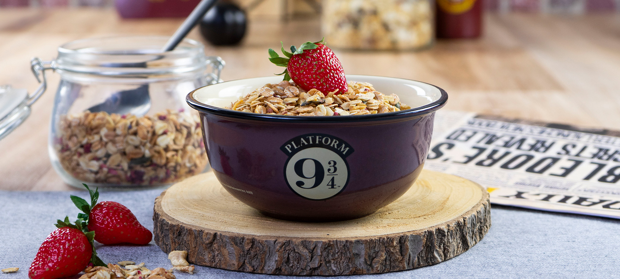 Eine Schale mit einem "Platform 9 3/4"-Logo enthält Müsli und ist mit einer Erdbeere gekrönt. Die Schale steht auf einer Holzplatte, im Hintergrund befinden sich noch mehr Müsli, Erdbeeren und ein Glas mit Müsli.
