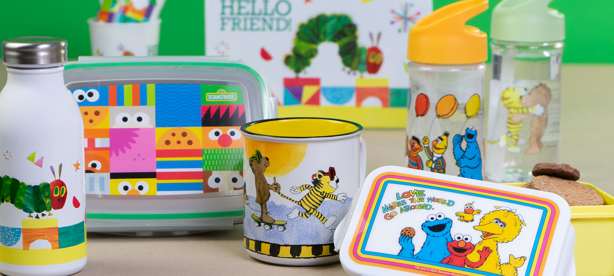 Ein farbenfrohes Sortiment von Lunch-Behältern, Flaschen und Bechern für Kinder mit Figuren wie der "Hungrigen Raupe Nimmersatt" und der "Sesamstraße" auf einer beigen Oberfläche mit grünem Hintergrund.