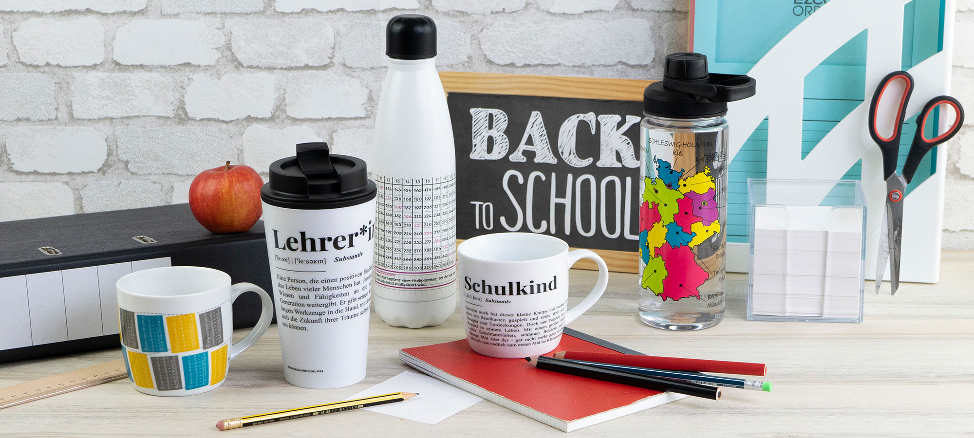 Ein Schreibtisch mit Schulsachen wie Bechern, Wasserflaschen, Notizbüchern, Stiften, Scheren, einem Apfel und einem Kreideschild mit der Aufschrift "Back to School" vor einer weißen Backsteinwand.