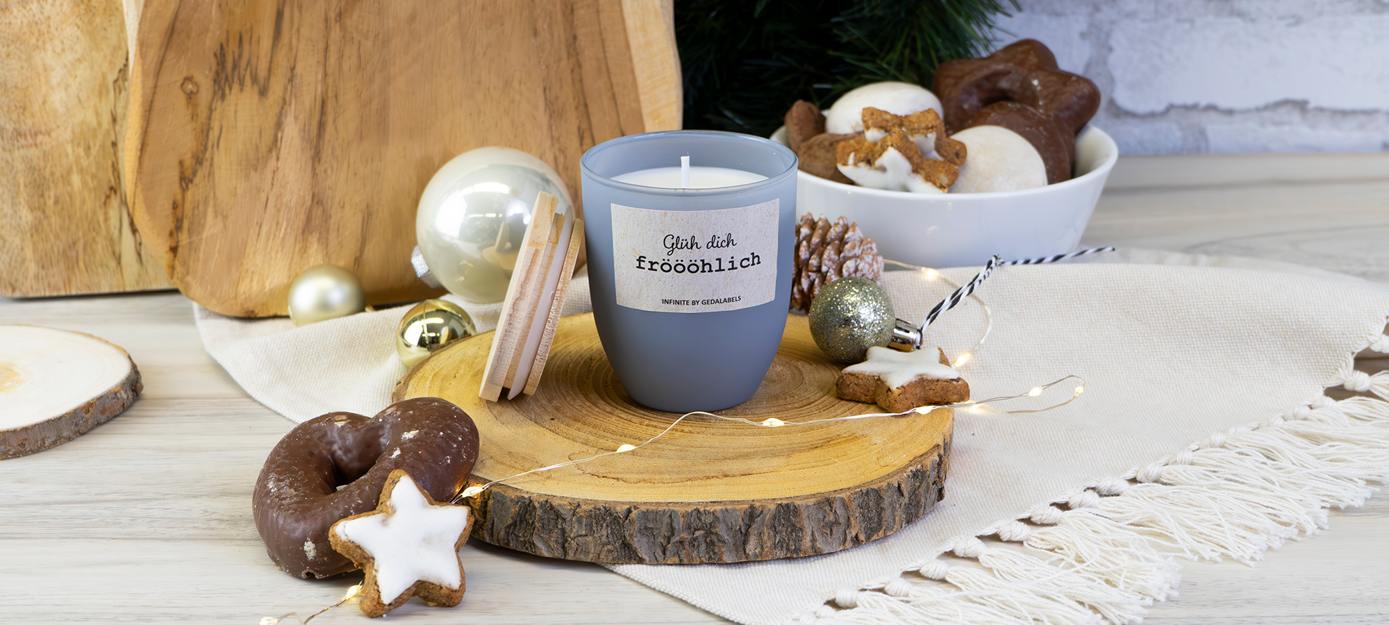 Eine graue Kerze auf einem Holztablett ist auf einem festlich gedeckten Tisch von Weihnachtsplätzchen, Ornamenten, Tannenzapfen und einer Schale mit Leckereien umgeben. Auf dem Kerzenetikett steht Gib dich fröhlich. Warme, gemütliche Weihnachtsatmosphäre.