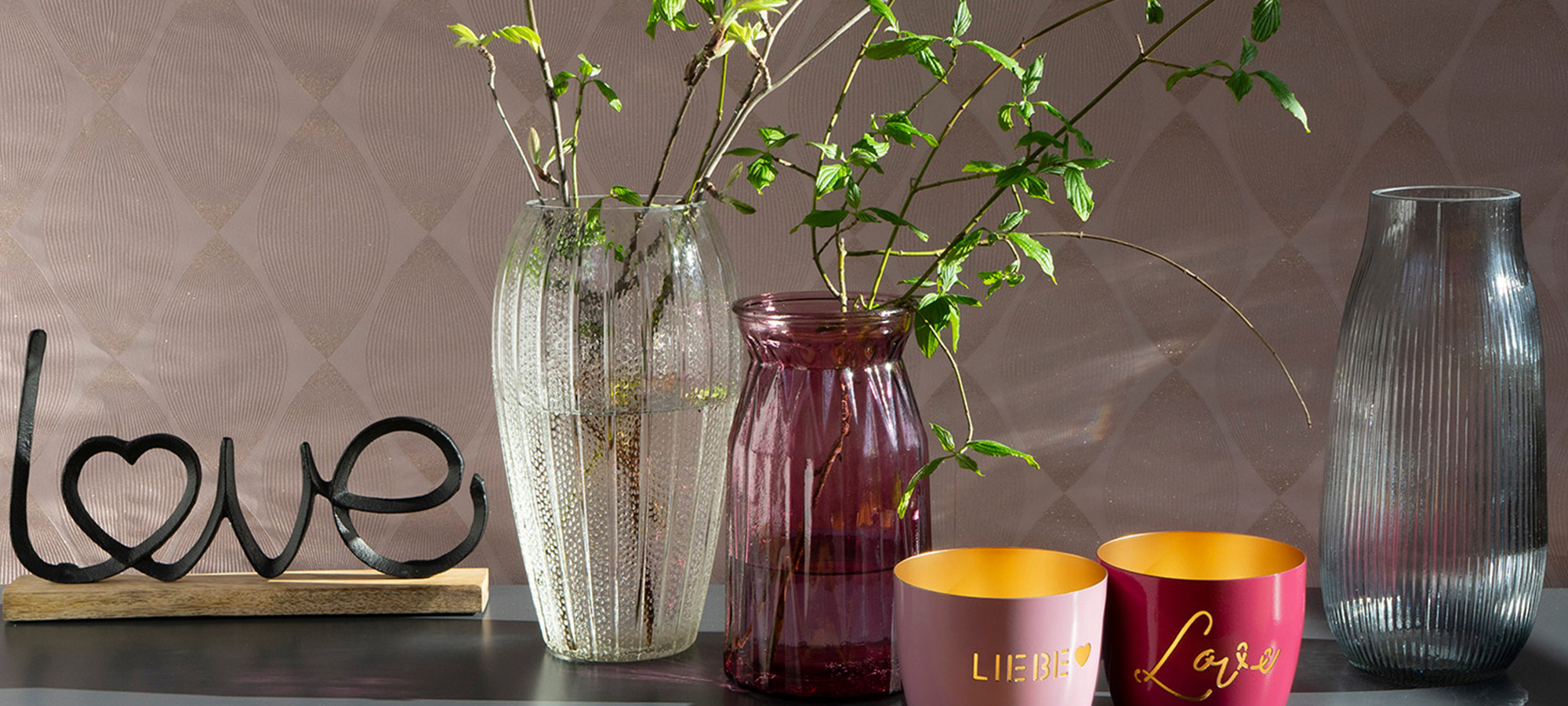 Ein dekoratives Arrangement mit Vasen mit grünen Zweigen, zwei roten und rosafarbenen Kerzen mit der Aufschrift LIEBE und LOVE, einer gerippten Glasvase und einem schwarzen Liebeszeichen aus Metall auf einem Holzsockel vor einer gemusterten Wand.