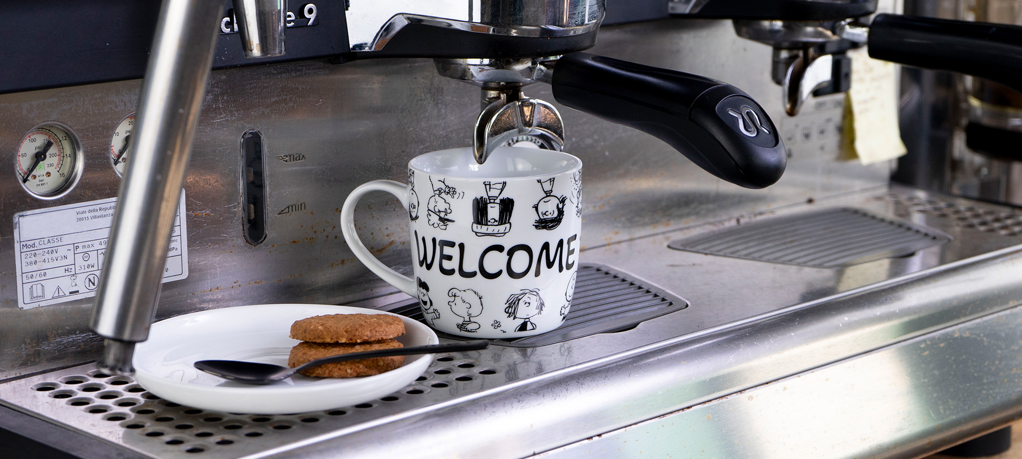 Eine Kaffeemaschine gießt Espresso in eine weiße Tasse mit WELCOME und Cartoon-Gesichtern. Daneben steht ein weißer Teller mit zwei Keksen und einem Löffel. Die Szene steht auf einem Metalltresen.