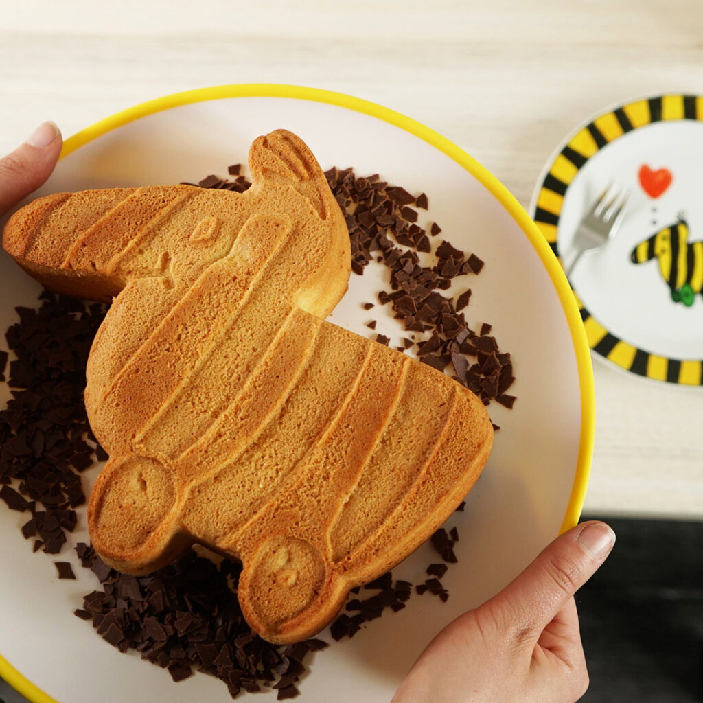 Eine Person hält einen Teller mit einem Kuchen in Form eines Hasen, umgeben von Schokoladenraspeln. Im Hintergrund sind ein Teller mit einem Bienenmotiv, eine Gabel und ein Löffel zu sehen.