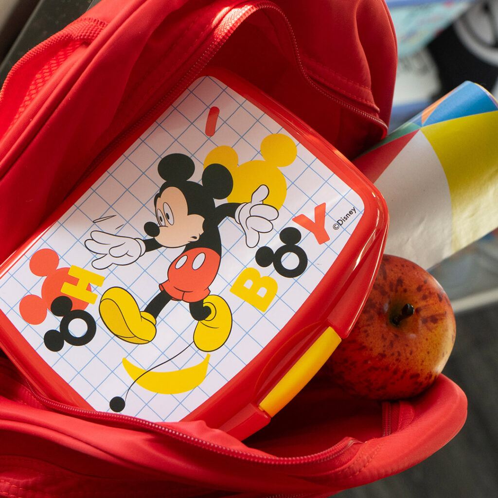 Ein roter Rucksack enthält eine Brotdose mit einem Micky-Maus-Motiv und der Aufschrift HI BOY auf einem gitterförmigen Hintergrund, einen Apfel und ein aufgerolltes buntes Papier.