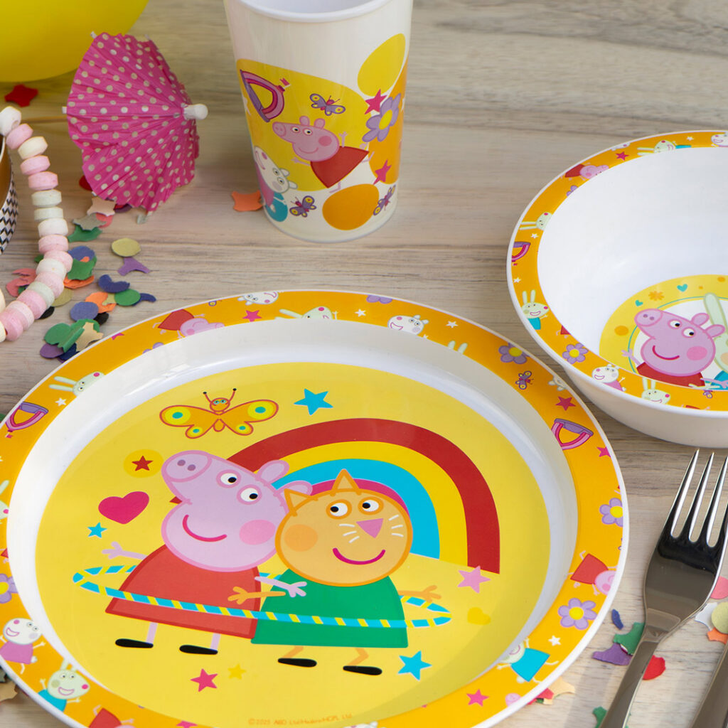 Kindergeschirr mit farbenfrohen Bildern von Peppa Pig und einer Katzenfigur mit einem Regenbogen, Blumen und Schmetterlingen auf gelben Tellern, einer Schüssel und einer Tasse, angeordnet auf einem Holztisch mit Konfetti und Partydekoration.