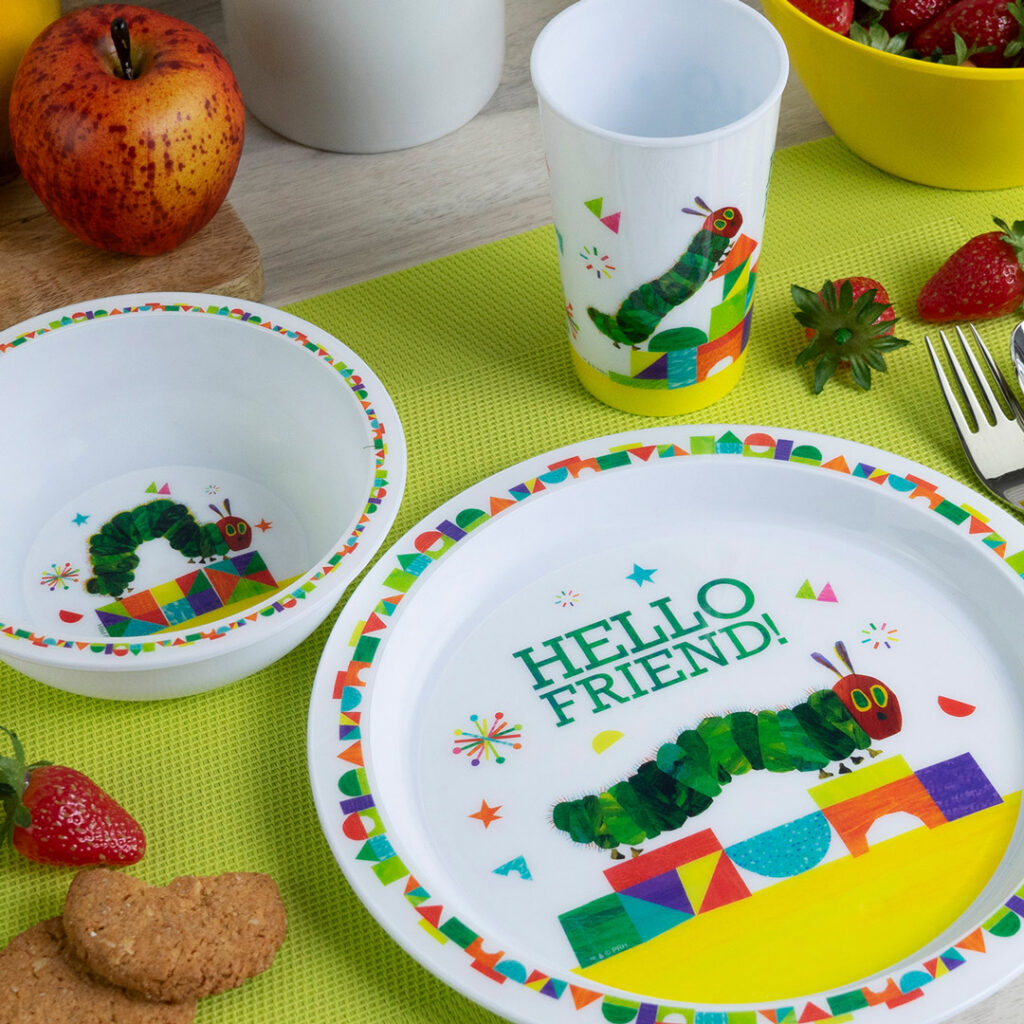 Das farbenfrohe Kindergeschirr-Set mit dem Motiv "The Very Hungry Caterpillar" besteht aus einer Schüssel, einem Teller mit der Aufschrift HELLO FRIEND! und einer Tasse auf einer grünen Matte, umgeben von Erdbeeren, Keksen, einem Apfel und Utensilien.