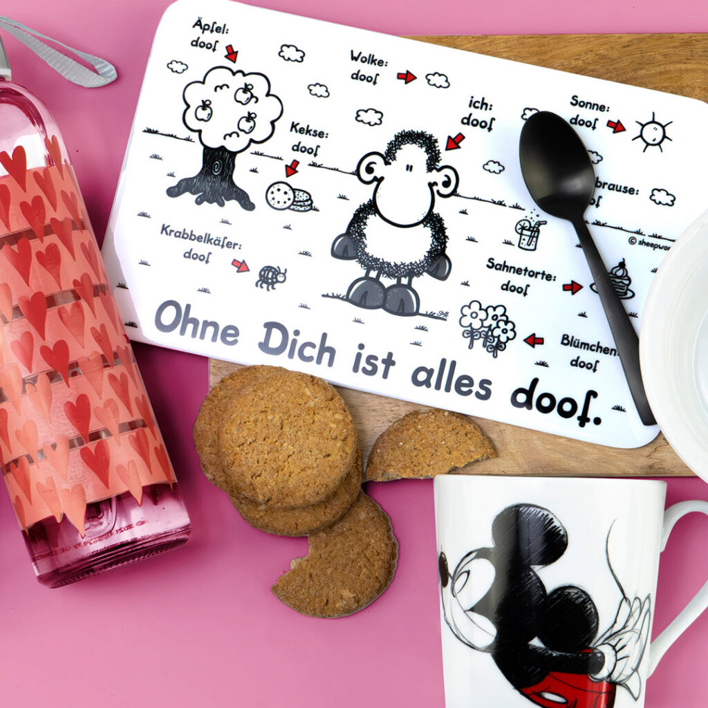 Ein deutsches Tischset mit der Aufschrift Ohne Dich ist alles doof! mit einem Cartoon-Schaf und verschiedenen beschrifteten Objekten. Drumherum stehen eine Tasse mit Mickey Mouse, Kekse, eine rosa Flasche mit Herzen und ein schwarzer Löffel auf einer rosa Fläche.