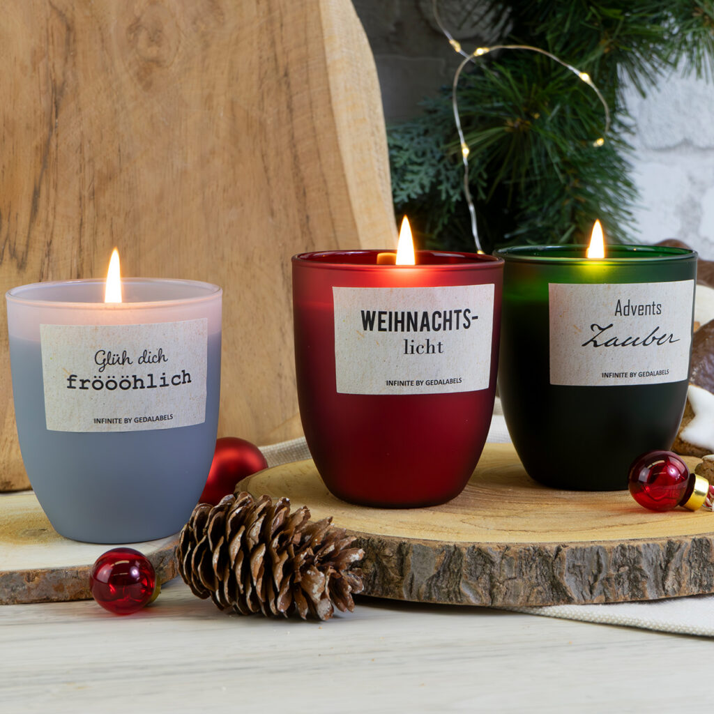 Drei brennende Weihnachtskerzen in Milchglashaltern - eine lavendelfarbene, eine rote und eine grüne - stehen auf einem Holzbrett mit festlichen Dekorationen, Tannenzapfen, rotem Schmuck und Grünzeug im Hintergrund.