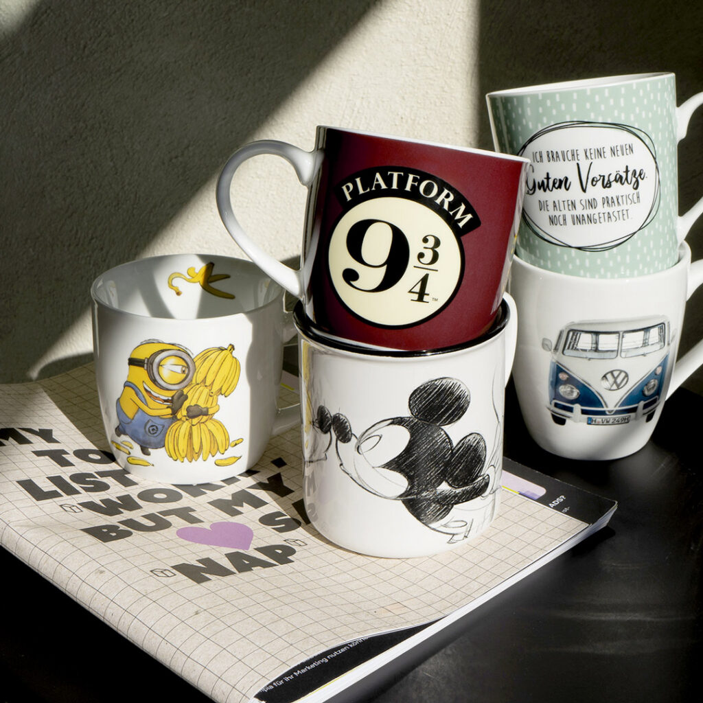 Fünf illustrierte Tassen sind über einem offenen Notizbuch mit Text gestapelt. Die Tassen zeigen Motive wie einen Minion, Harry Potters Bahnsteig 9¾, Mickey Mouse, handgeschriebene Zitate und einen blauen VW-Bus. Das Sonnenlicht scheint auf die Szene.