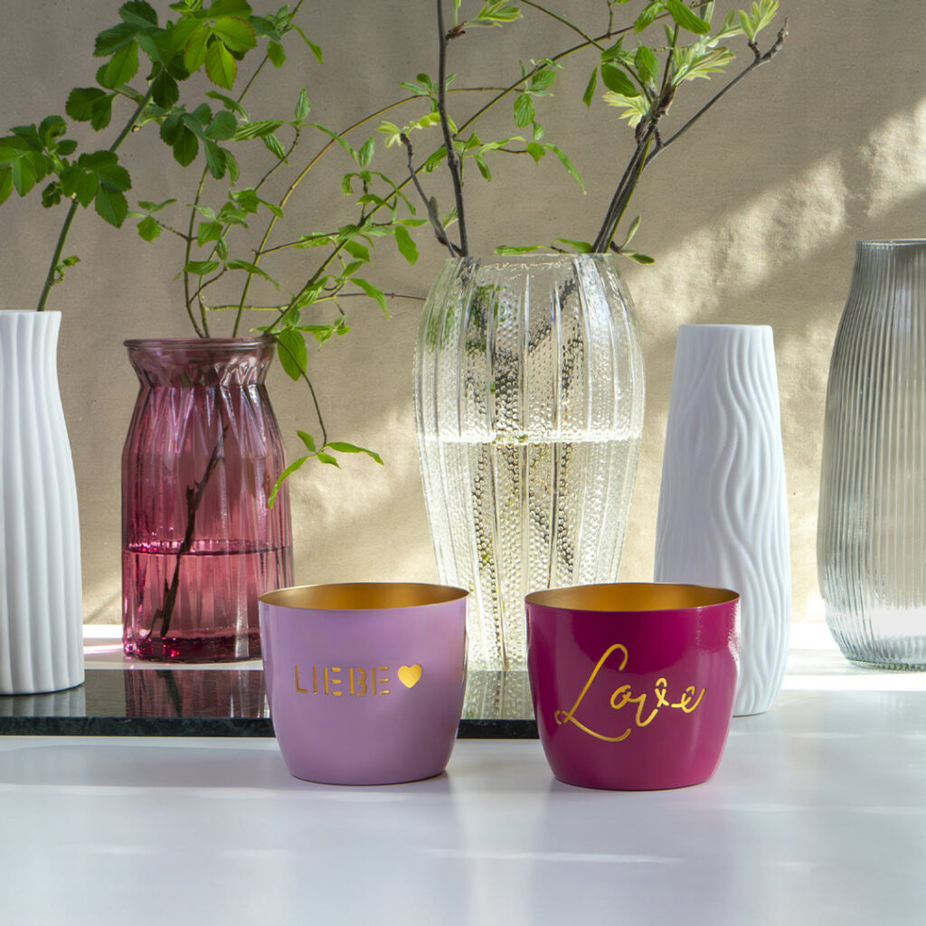 Auf einer weißen Fläche stehen zwei rosafarbene Kerzenständer mit goldenem Innenleben, einer mit der Aufschrift LIEBE und einem Herz, der andere mit Love. Dahinter stehen dekorative Glas- und Keramikvasen mit grünen Blätterzweigen.
