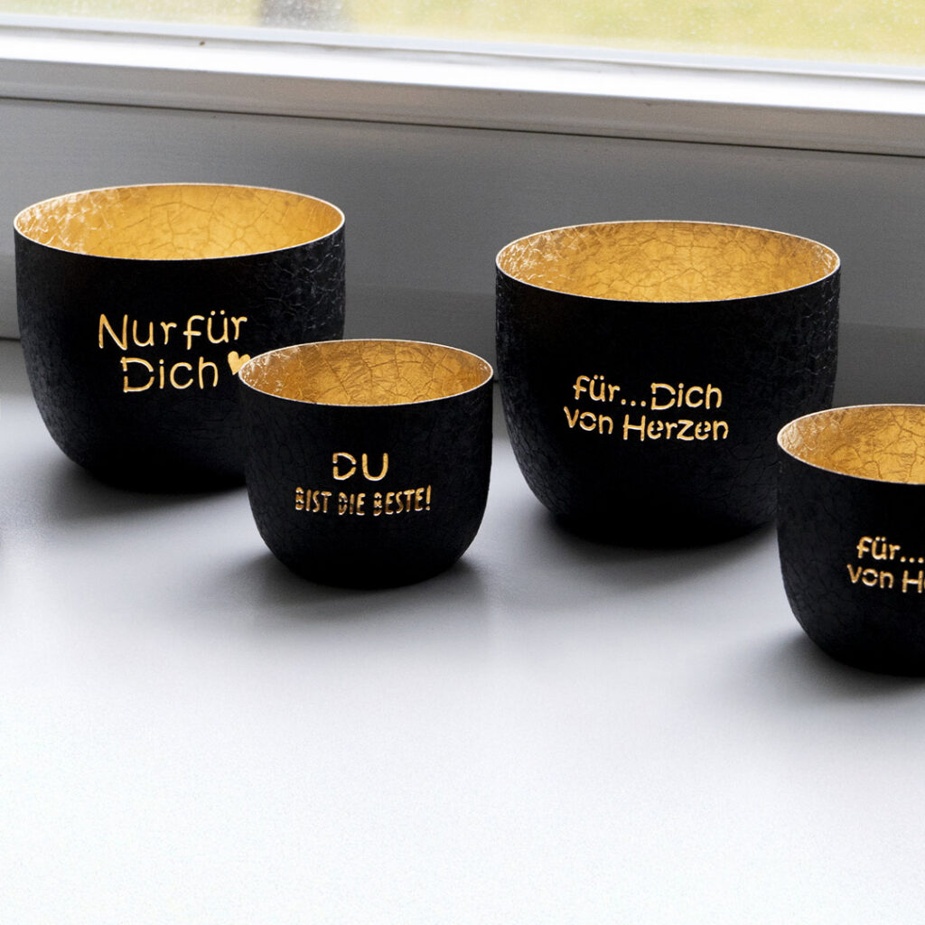 Vier schwarz-goldene Tassen mit deutschem Text sind auf einer weißen Fläche neben einem Fenster angeordnet. Die Texte enthalten Sätze wie "Nur für Dich" und "Du bist die Beste!" in goldenen Lettern.