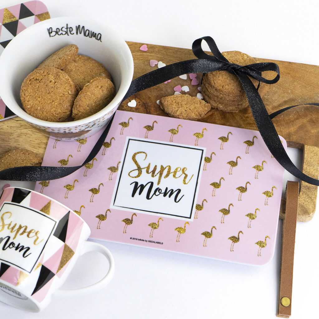 Ein rosafarbenes Tischset mit goldenen Flamingos und dem Text Super Mom, eine Tasse und eine Schüssel mit Keksen, ein Holzbrett und ein schwarzes Band. Auf der Schale steht Beste Mama. Die Szene ist hell und festlich.