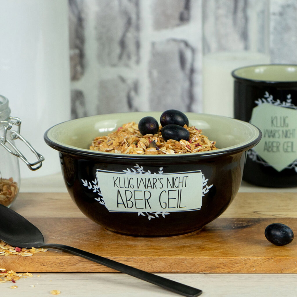 Eine schwarze Keramikschüssel, gefüllt mit Müsli und bestückt mit Weintrauben, steht auf einem Holzbrett. Die Schale ist mit "Klug wars nicht aber geil" beschriftet. Im Hintergrund stehen ein passender Becher, ein Glasgefäß und eine Kerze.