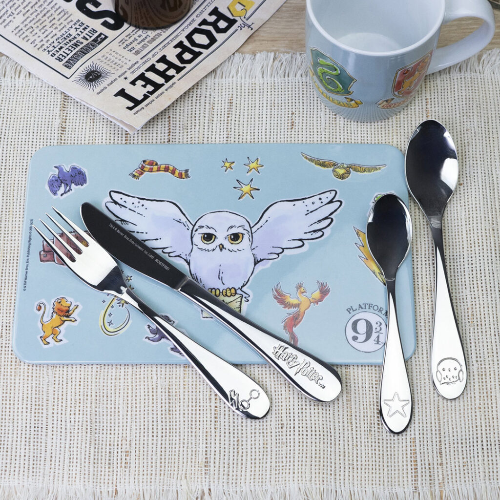 Ein Tischset mit Illustrationen zum Thema Harry Potter, darunter die Eule Hedwig, wird zusammen mit einer Tasse und vier Besteckteilen, die jeweils mit Harry-Potter-Motiven graviert sind, auf einer gewebten Matte neben einer gefalteten Zeitung aufgestellt.