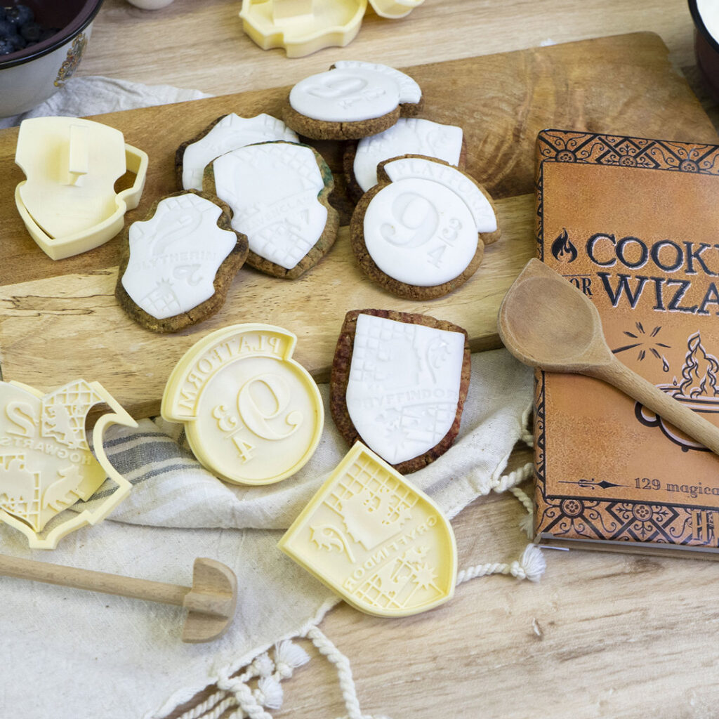 Ein Holztisch mit Keksen in Form von Schilden, verziert mit weißem Zuckerguss, auf dem die Namen der Häuser von Hogwarts und die Zahl 9 stehen. In der Nähe liegen Keksstempel, ein Holzlöffel und ein Kochbuch mit dem Titel Kochen für Zauberer.