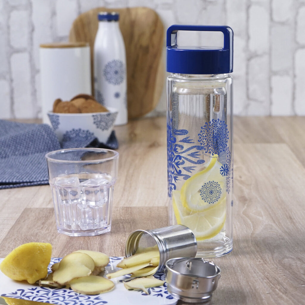Eine gläserne Wasserflasche mit blauen Mustern, gefüllt mit Wasser, Zitronenscheiben und Ingwer, steht auf einem Holztisch mit einer passenden Tasse, Ingwerstücken, Keksen und einer dekorierten Flasche im Hintergrund.