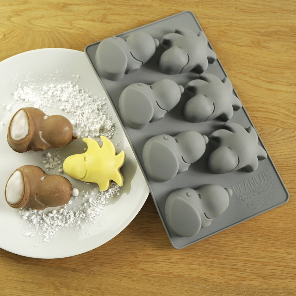 Eine graue Silikonform mit den Gesichtern der Peanuts-Figuren, daneben ein weißer Teller mit gelbem und braunem Gebäck in Form der Figuren, bestäubt mit Puderzucker.