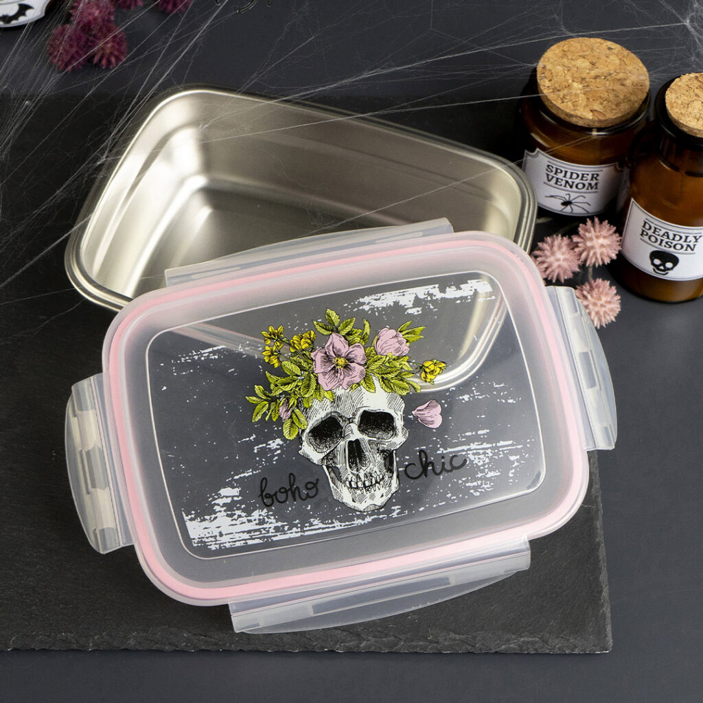 Ein rechteckiger Lebensmittelbehälter aus Kunststoff mit einem durchsichtigen Deckel, auf dem ein Totenkopf mit Blumen und der Aufschrift "Boho Chic" abgebildet ist. In der Nähe befinden sich ein Metalltablett, zwei braune Flaschen mit der Aufschrift "Spinnengift" und "tödliches Gift" sowie lila Dekorationen.