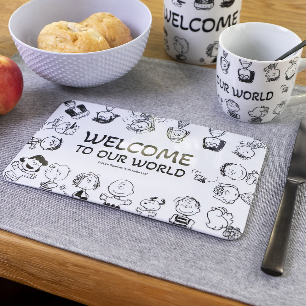 Ein Tischset mit Peanuts-Figuren und dem Text WELCOME TO OUR WORLD liegt auf einem Tisch mit einem Messer, einer Tasse, einer Schüssel Brot und einem Apfel auf einer grauen Stoffserviette.