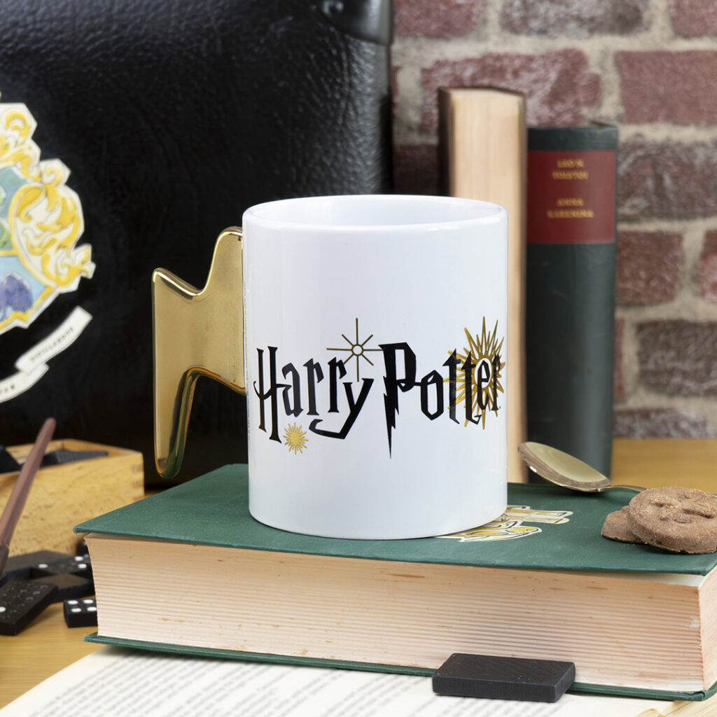 Ein weißer Becher mit dem Harry-Potter-Logo und einem goldenen Blitzgriff steht auf einem geschlossenen Buch, umgeben von Keksen, Büchern, einem Löffel und anderen Harry-Potter-Gegenständen auf einem Holztisch.