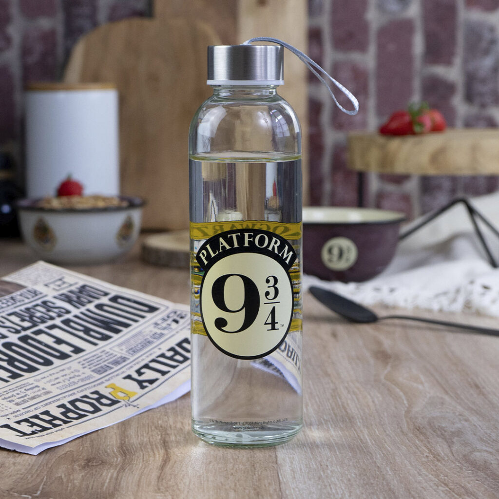 Eine Klarglas-Wasserflasche mit einem silbernen Deckel und einem Etikett der Plattform 9¾ steht auf einem Holztisch. Im Hintergrund liegen eine Zeitung, eine Schüssel, ein Schneidebrett und einige Erdbeeren.