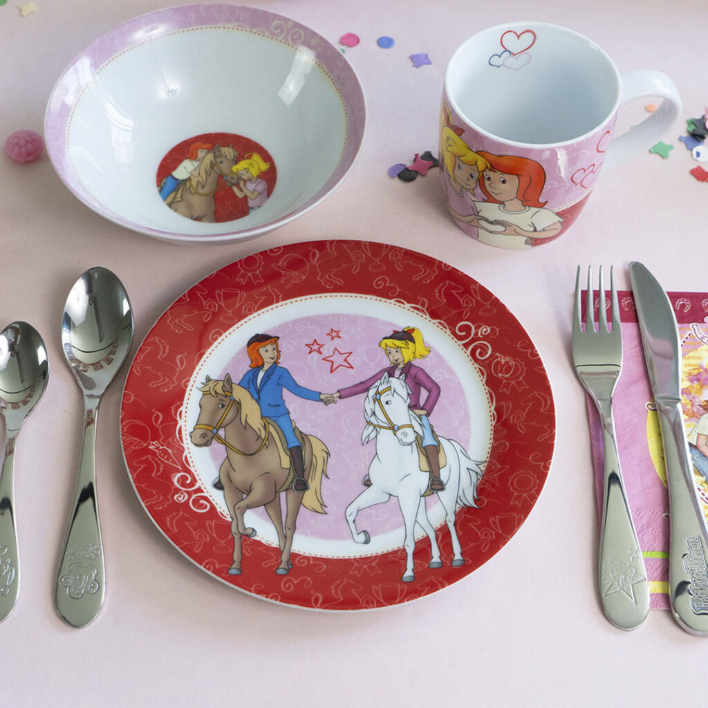 Kindergeschirrset mit Teller, Schüssel und Becher mit Prinzessinnen auf Pferden, Gabel, Messer, Löffel und Serviette auf einer rosa Fläche, die mit Konfetti und kleinen Dekoartikeln verziert ist.