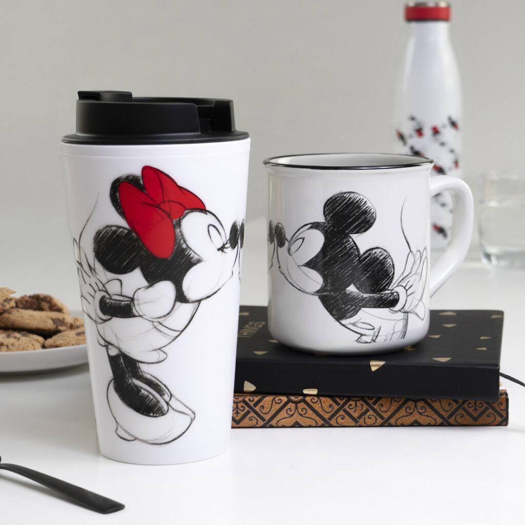 Zwei weiße Tassen mit Schwarz-Weiß-Skizzen von Minnie Mouse und Mickey Mouse, die sich für einen Kuss anlehnen, platziert auf einem dekorativen Notizbuch, mit Keksen, einer Wasserflasche und einem Glas im Hintergrund.