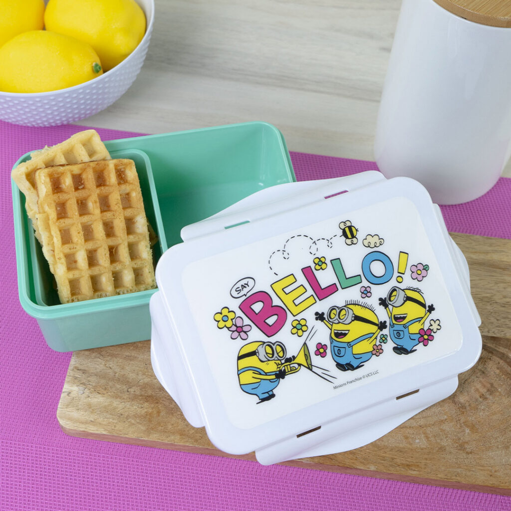 Eine weiße Brotdose mit einem Minions-Motiv und dem Wort BELLO! auf dem Deckel ist geöffnet und offenbart Waffeln in einem grünen Fach. Zitronen in einer Schüssel und ein weißer Behälter stehen in der Nähe auf einem Holzbrett und einer rosa Matte.