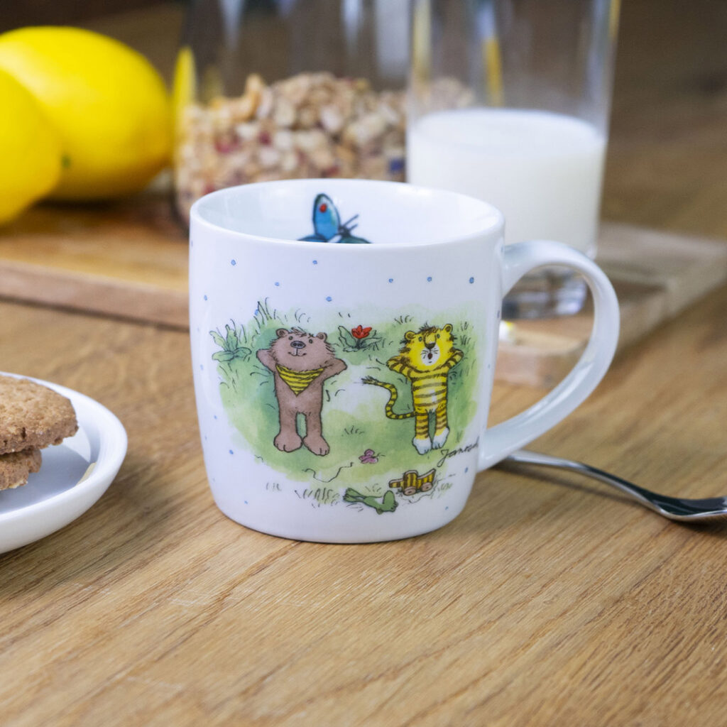 Ein weißer Becher mit bunten Abbildungen eines Bären und eines Tigers steht auf einem Holztisch. In der Nähe stehen ein Glas Milch, Zitronen, eine Schüssel mit Müsli und ein weißer Teller mit Keksen.