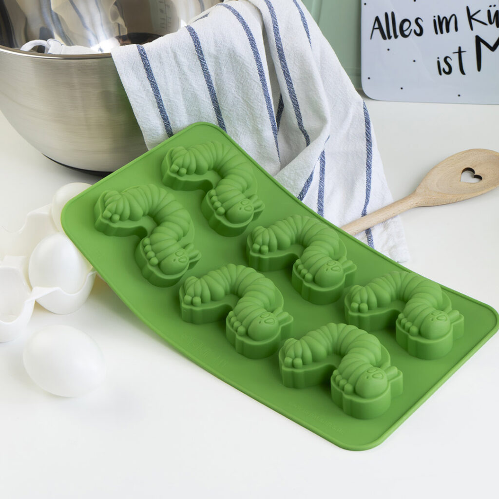 Eine grüne Silikonbackform mit raupenförmigen Vertiefungen steht auf einem Küchentisch in der Nähe von Eiern, einem Holzlöffel mit einem Herzausschnitt, einem gestreiften Handtuch und einer Metallschüssel.
