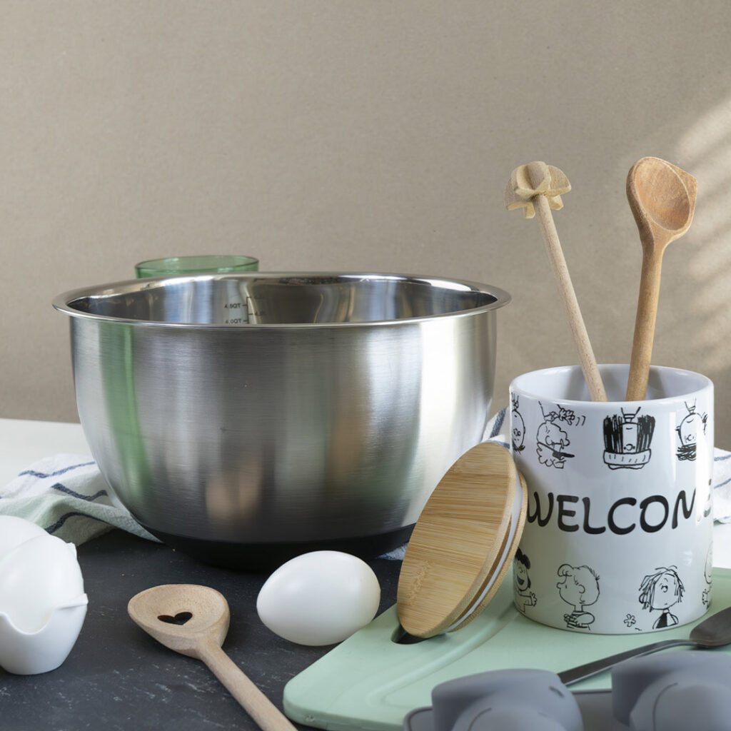 Auf dem Tresen steht eine Metallschüssel neben einem Becher mit der Aufschrift WELCOME, in dem sich Holzlöffel, ein ganzes Ei, ein Eierhalter, ein grünes Schneidebrett und ein gestreiftes Handtuch befinden.