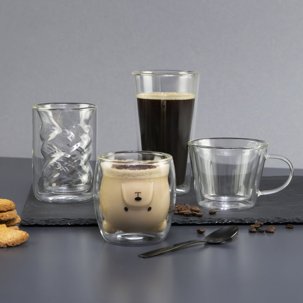 Vier verschiedene Becher aus klarem Glas, darunter einer mit einem niedlichen Bärengesicht und Ohren, gefüllt mit einem cremigen Getränk, werden zusammen mit Keksen, Kaffeebohnen und einem Löffel auf einer dunklen Oberfläche und einem Schiefertablett präsentiert.