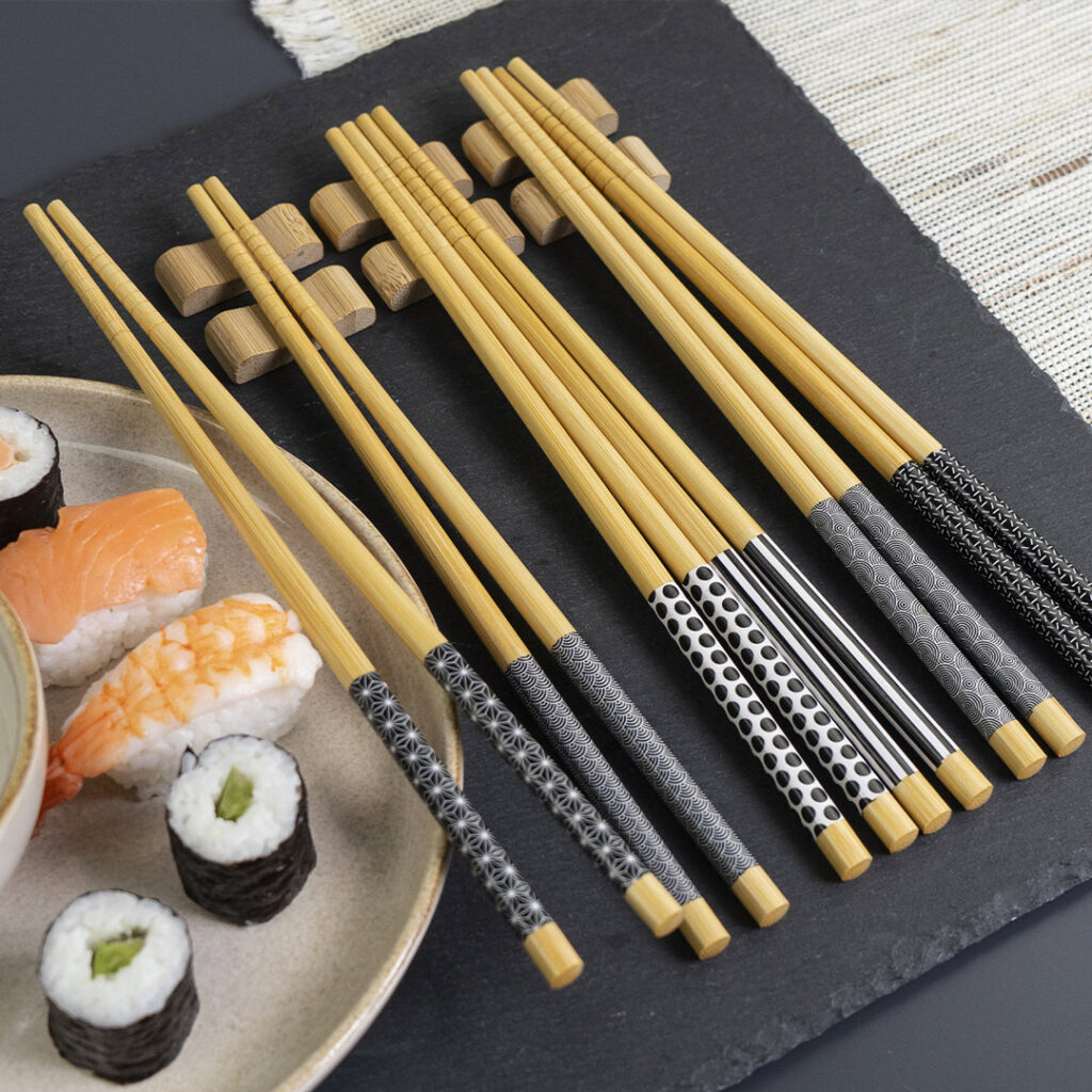 Ein Satz Bambus-Essstäbchen mit gemusterten schwarz-weißen Griffen liegt auf einer Schieferplatte neben einem Teller mit verschiedenen Sushi-Stücken, darunter Nigiri und Maki-Rollen.