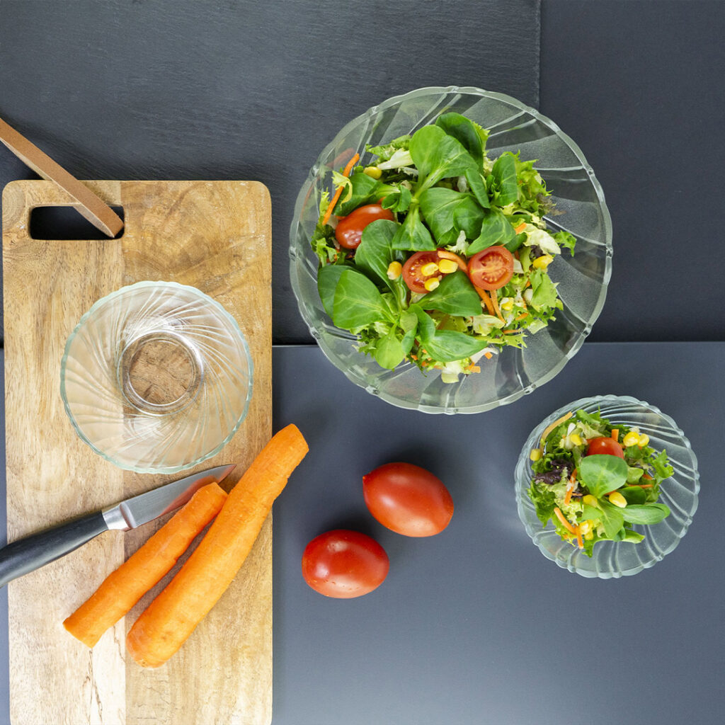 Draufsicht auf einen Salat in Glasschalen, mit zwei ganzen Tomaten, zwei Karotten, einem Messer und einer leeren kleinen Schüssel auf einem hölzernen Schneidebrett auf einer dunklen Fläche.